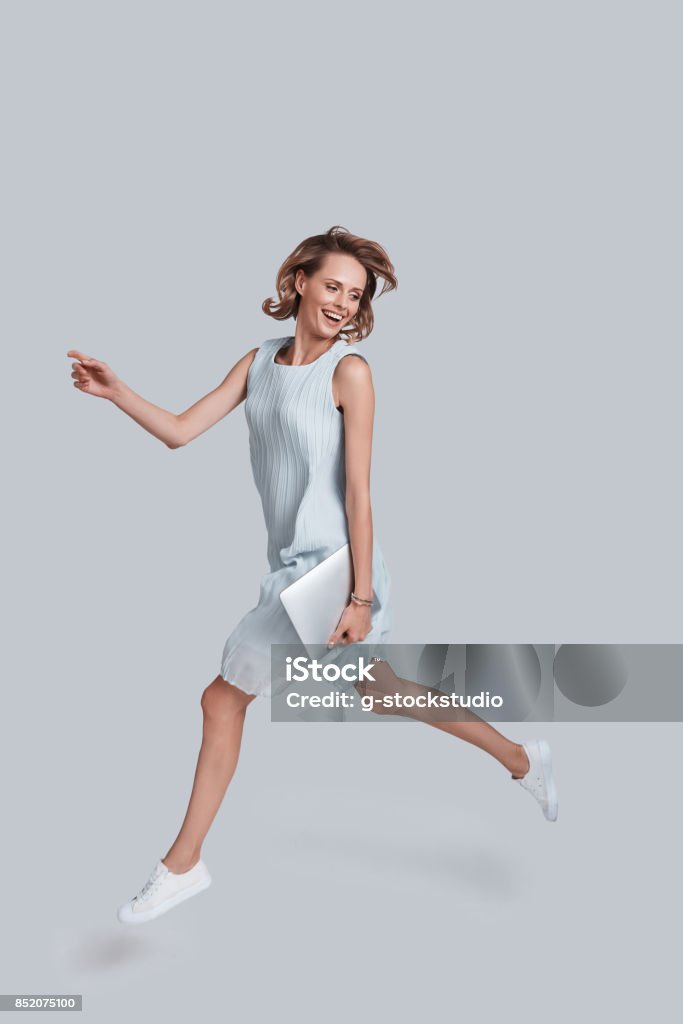 Mid-air motion. Full length of playful young woman gesturing and smiling while jumping against grey background Women Stock Photo