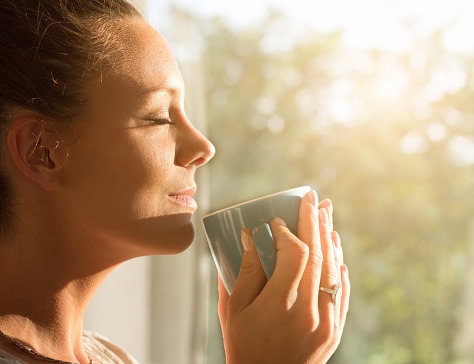 Pretty young woman enjoying first morning coffee on sunshine by window