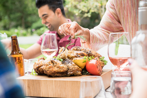 Cropped view of food on dining table and wine glasses, woman's hand holding fresh rosemary, cooked meat on wooden chopping board close up