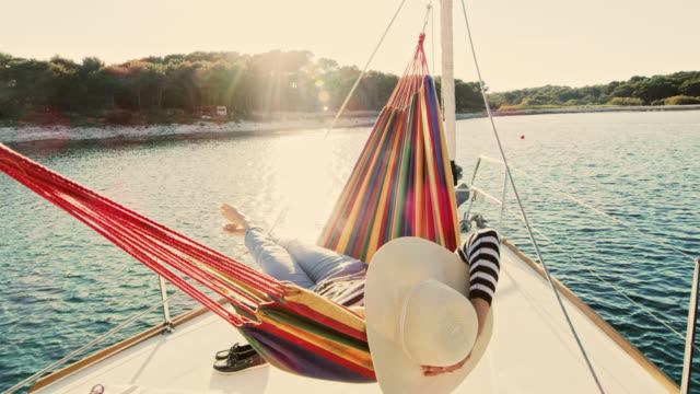 Slow motion wide shot of a woman with a sunhat lying in a hammock on a sailboat deck. Shot was taken in the late afternoon sun. Shoot in 8K resolution.