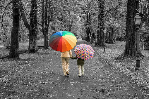 Rear view on mother and son with multi colored umbrellas in isolated color walking hand in hand in black and white park. Maybe useful as a symbol for guidance and security in childhood.