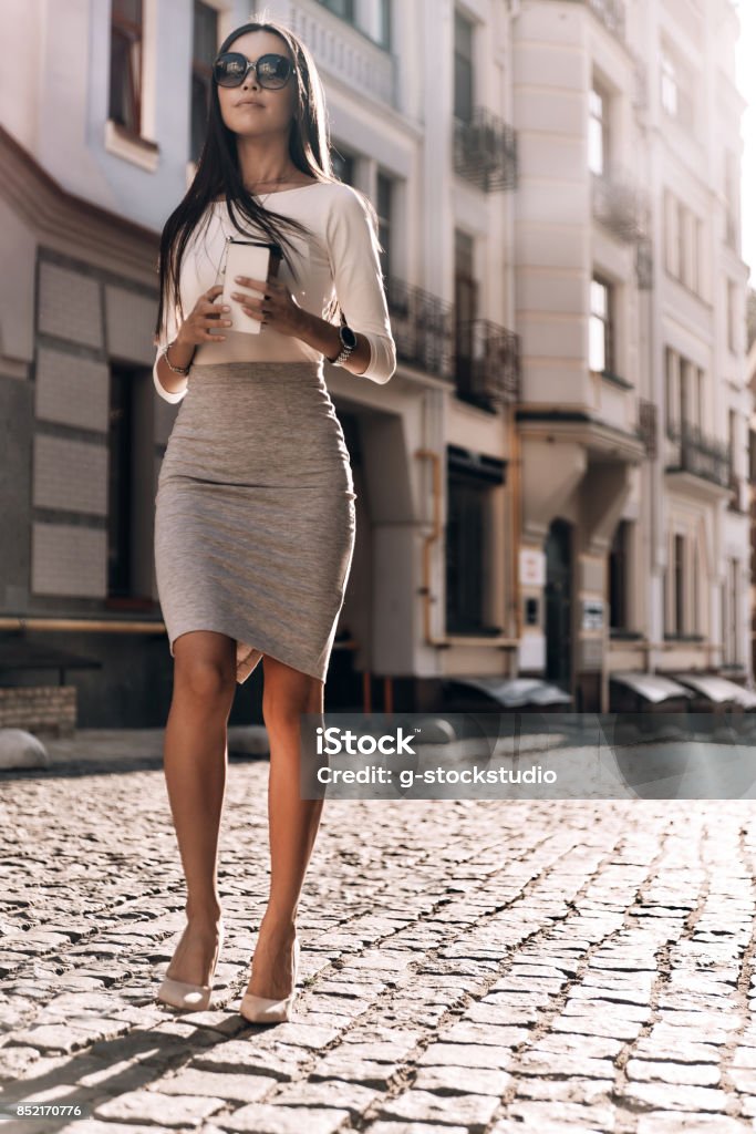 Enjoying her coffee break. Full length of attractive young woman holding her smart phone and disposable cup while walking outdoors Businesswoman Stock Photo