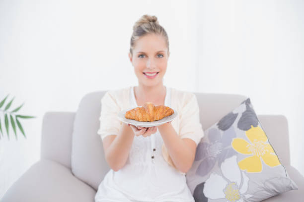 Cheerful fresh model holding croissant sitting on sofa in bright living room