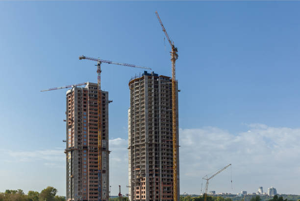 Several modern high-rise buildings in different construction stages with yellow cranes and unfinished concrete base under gray sky, concept of architecture