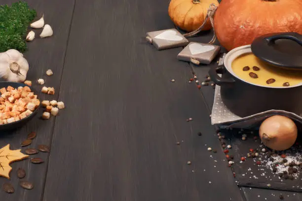 Pumpkin creme soup in a dark ceramic pan on the dark background. Ingredients: pumpkins, carrots, onions, garlic, parsley, salt and pepper, also croutons and pumpkin seeds for serving . Text space.