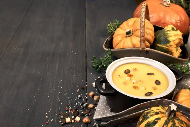 Pumpkin creme soup in a dark ceramic pan on the dark background. Ingredients: pumpkins, onions, parsley, salt and pepper, also croutons and pumpkin seeds for serving . Text space.