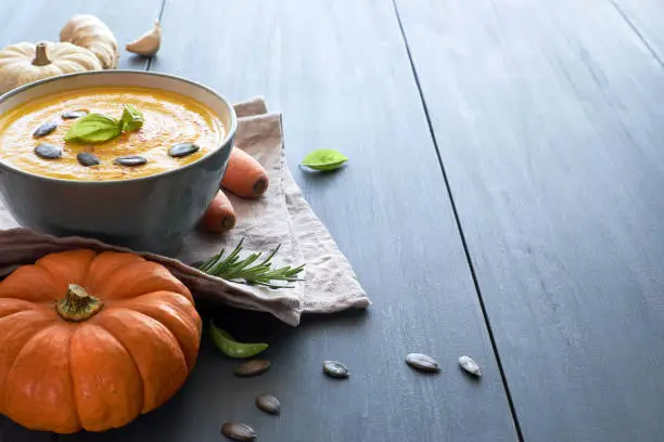 Pumpkin creme soup with carrot, ginger and garlic, served with pumpkin seeds and basilicum. Ingredients: pumpkin, carrot, ginger, onion and garlic. The soup is in ceramic bowl on blue wood, text space.
