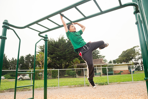 This is a royalty free, color photograph of a young California Bay area, mixed race boy spending time playing outdoors in the park. He wears a green shirt as he swings from the monkey bars. Photographed with a Nikon D800 DSLR camera.