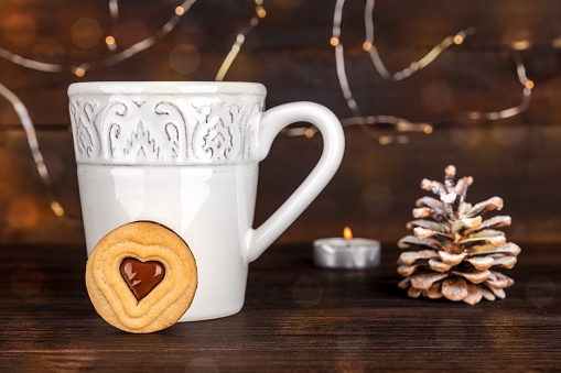 A photo of a cookie with a cup of hot chocolate behind it, with a snowy pine cone, a candle, and fairy lights in the blurred background, with a place for text, side view, on dark textures