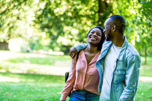 Couple walking around the bounds of a lush green park, with the man's arm around his girlfriend.