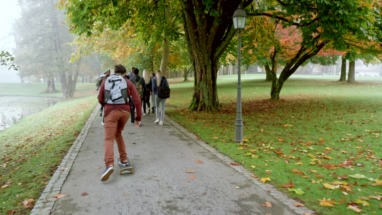 Caucasian male student riding his skateboard past a group of student walking through a park on a fall morning