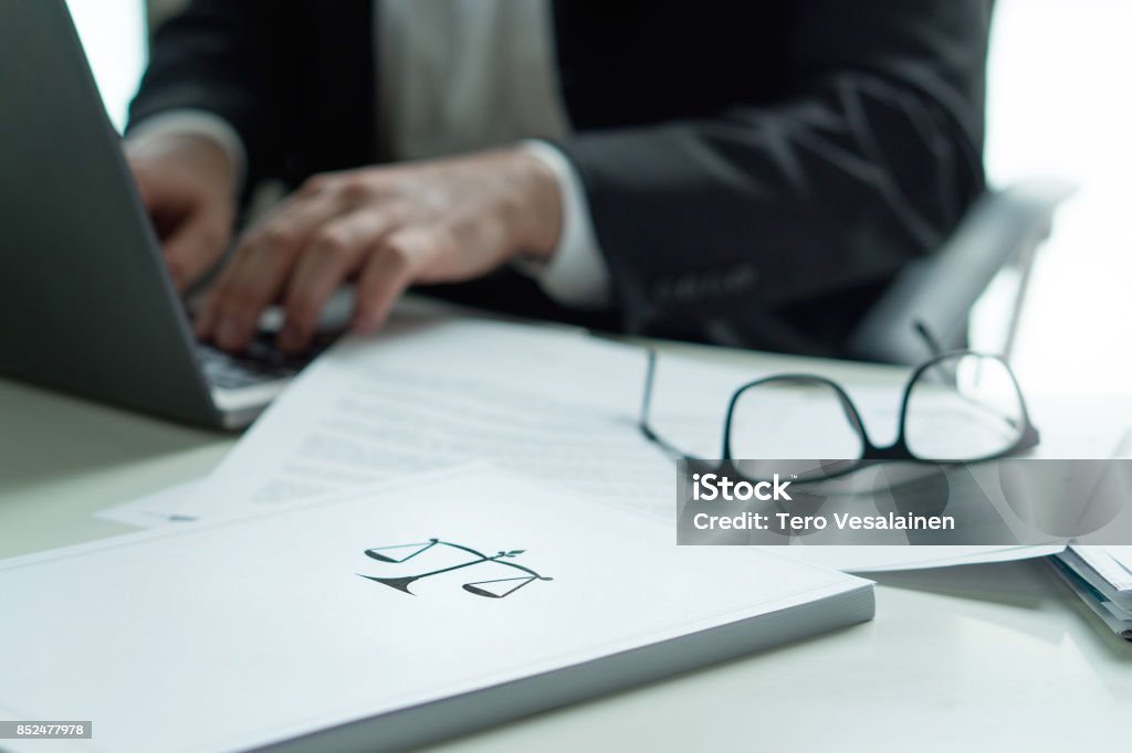 Lawyer working in office. Attorney writing a legal document with laptop computer. Glasses on table. Pile of paper with scale and justice symbol. Law firm and business concept. Lawyer working in office. Attorney writing a legal document with laptop computer. Glasses on table. Pile of paper with scale and justice symbol. Lawyer Stock Photo
