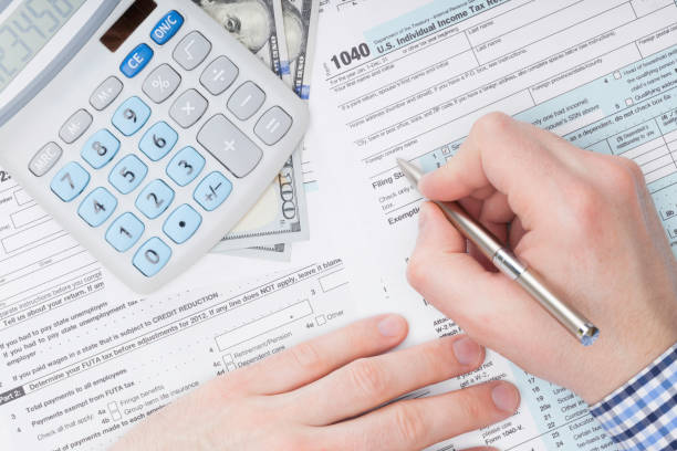 Close up shot of a male filling out US 1040 Tax Form next to calculator and 100 dollars banknote under it stock photo