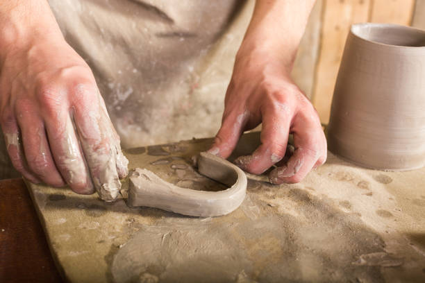 istock potter, stoneware, ceramics art concept - craftsman fingers forming the handle from raw fireclay, master hands working with some piece of clay, male stand at a workshop near the table 852617456