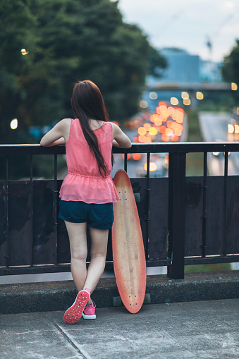 A young Japanese woman is with a skateboard watching the city at dusk.