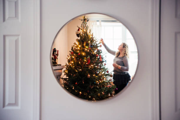 Mature woman ldecorating Christmas tree at home. She is in the living room, in front of a window. This was shot in a round mirror in the back of the room. Horizontal indoors mid length shot with copy space. This was taken in Quebec, Canada.