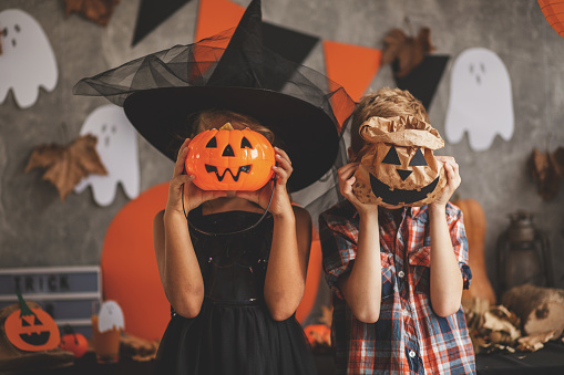 Boy and a girl are playing at the Halloween party, covering their faces with Jack O'Lantern bucket and paper bag.