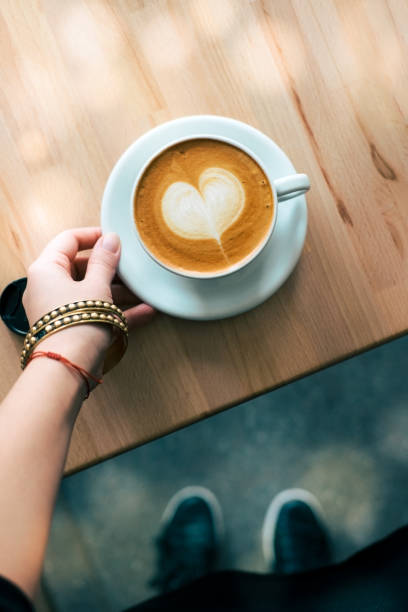 Woman hand holding white mug of cappuccino coffee with heart shaped latte art. Wooden table. V iew with feet from above.