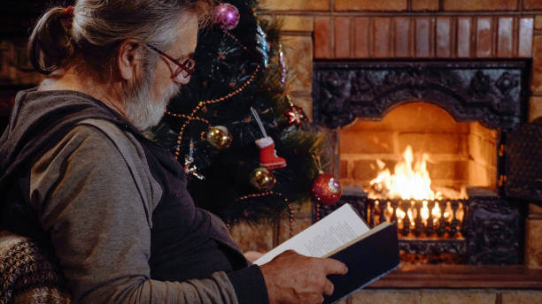 Senior man reading book near the fireplace and Christmas tree at xmas eve stock photo
