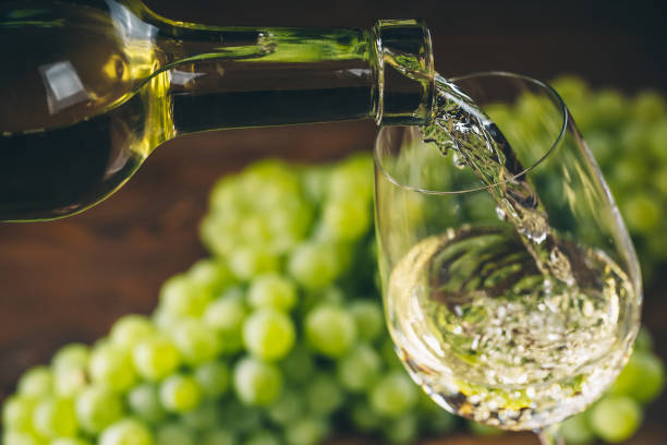 Pouring white wine into a glass with a bunch of green grapes against wooden background stock photo
