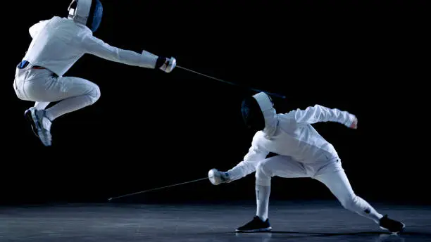 Two Professional Fencers Show Masterful Swordsmanship in their Foil Fight. They Dodge, Leap and Thrust and Lunge. Shot Isolated on Black Background.