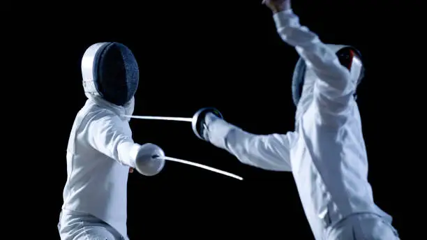 Two Professional Fencers Show Masterful Swordsmanship in their Foil Fight. They Attack, Defend, Leap and Thrust and Lunge. Shot Isolated on Black Background.
