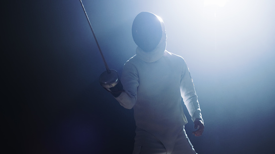 Fully Equipped Fencer Puts Lifts Foil Sword in Readiness for a Match. He Stands in the Spotlight while Darkness is Around Him. Shot Isolated on Black Background.