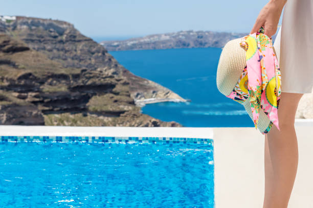 Horizontal color image of woman standing at the edge of terrace and holding sun hat. Aegean sea and landscape in the background.