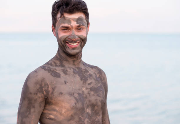 Portrait of handsome man with mineral mud of Dead Sea. Portrait of tourist man covered by the mineral mud of Dead Sea. The handsome man looking at camera with a friendly smile. Dead Sea minerals, health care, black mud, beauty skin treatment. people covered in mud stock pictures, royalty-free photos & images