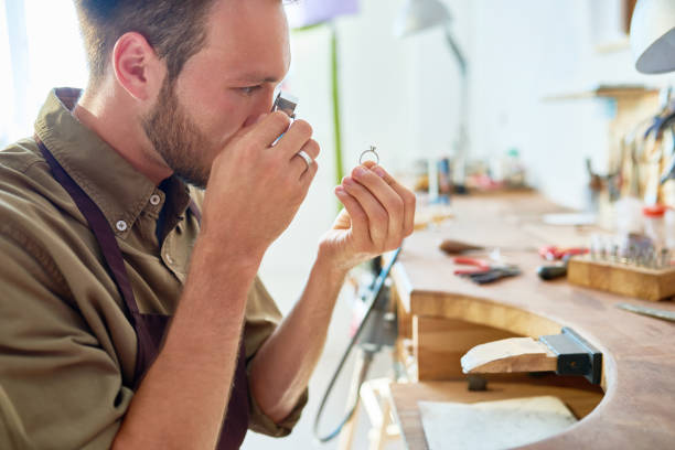 Young Jeweler Appraising Ring in Shop Side view portrait of jeweler inspecting ring through magnifying glass in workshop jewelry store stock pictures, royalty-free photos & images