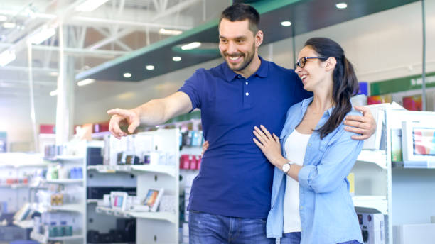 istock In the Electronics Store Beautiful Young Couple are Looking for New Electrical Device. Men Points his Finger at Something. Store is Big, Bright and Has all the New Devices. 853324106