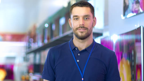 Portrait of a Professional Expert Consultant Smiles and Looks into Camera as Stands in the Bright, Modern Electronics Store Full of Latest Models of TV Sets, Cameras, Tablets and other Devices.The Depth of Field Shot. retrato de un profesional experto consultor sonrisas y miradas a cámara como soportes en la brillante, moderna electrónica tienda completo de últimos modelos de televisores, cámaras, tablets y otros devices.the profundidad de campo de tiro.
