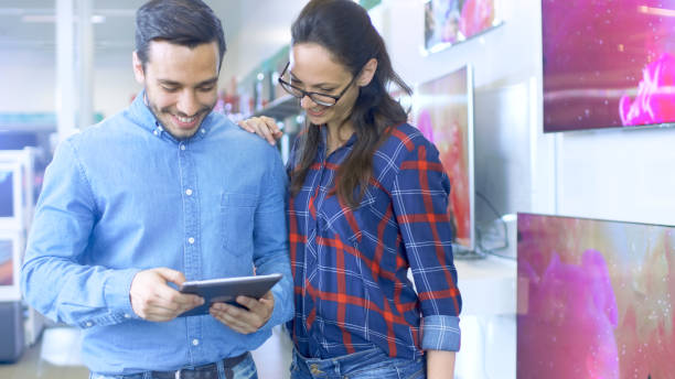 istock Young Beautiful Couple In the Electronics Store Looks at Latest Tablet Computer Models, Choosing Right One for Their Use and Budget. Store is Bright and Modern, Has all The Latest Devices. 853324168