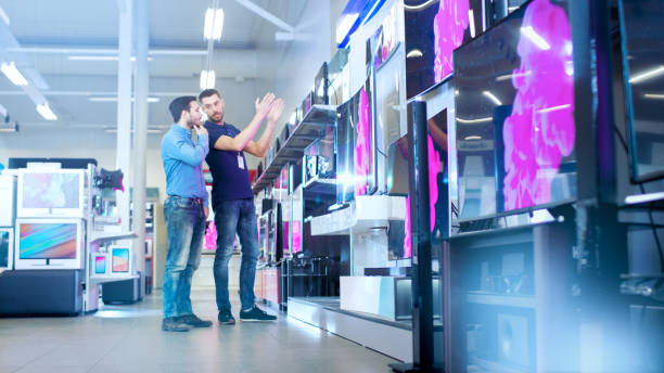 In the Electronics Store Professional Consultant Shows Latest 4K UHD TV's to a Young Man, They Talk about Specifications and What Model is Best for Young Man's Home. Store is Bright, Modern and Has all the Latest Models. en los televisores de uhd electrónica tienda profesional consultor muestra últimos 4k a un hombre joven, hablan sobre las especificaciones y el modelo de lo que es mejor para el hogar de la joven. tienda es brillante, moderna y tiene todos los últimos modelos.