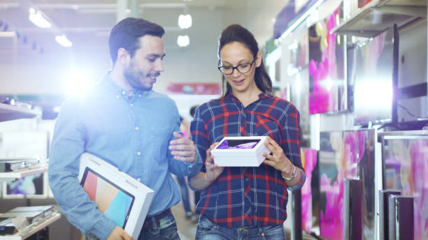 heureux couple marchant dans le magasin d’électronique, ils ont acheté le nouveau modèle de la tablette tactile pour lui et nouveau smartphone pour elle. centre commercial est grand, lumineux et complète de l’électronique plus récente. - ipad shopping gift retail photos et images de collection