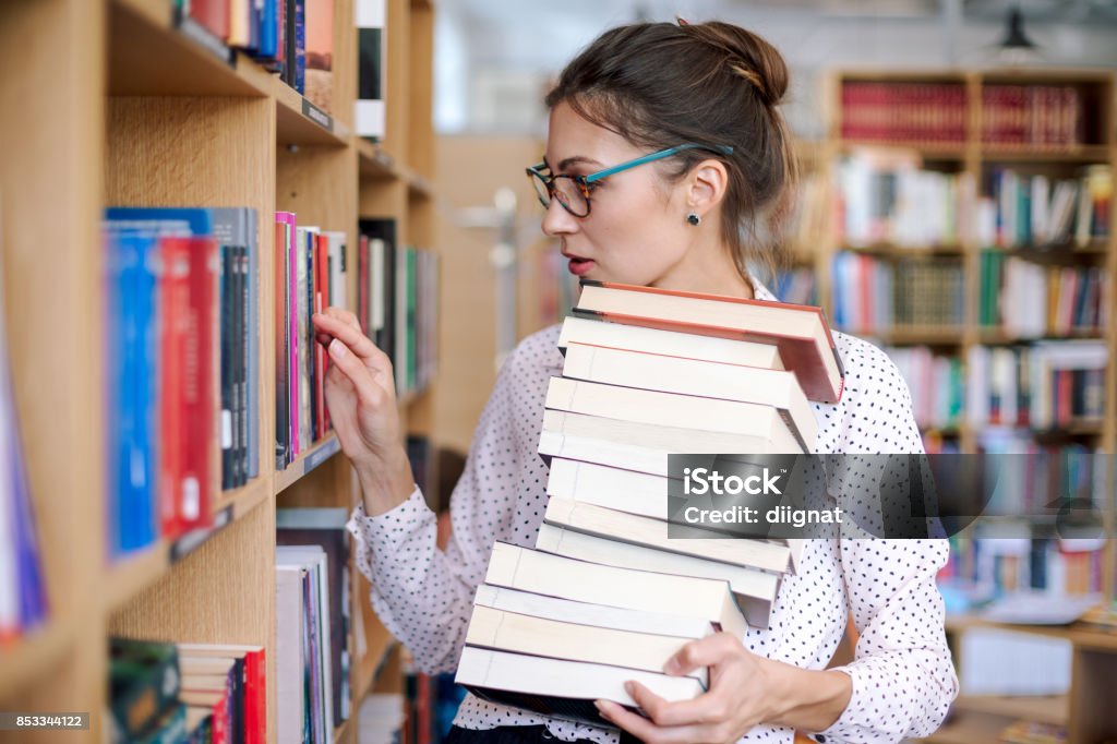 Young woman with a stack of books in library want more Attractive young woman wearing pink pale shirt with dots and fashionable glasses holding a stack of books in the library surrounded by bookshelves with colorful books. Book grabbing woman. Library Stock Photo