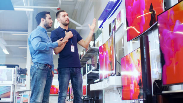 nel negozio di elettronica consulente professionale mostra le ultime tv uhd 4k a un giovane, parlano di specifiche e quale modello è meglio per la casa di un giovane. il negozio è luminoso, moderno e ha tutti gli ultimi modelli. - retail shopping talking customer foto e immagini stock