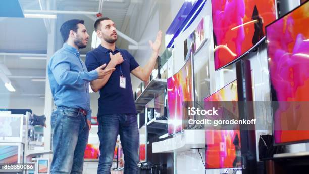 In The Electronics Store Professional Consultant Shows Latest 4k Uhd Tvs To A Young Man They Talk About Specifications And What Model Is Best For Young Mans Home Store Is Bright Modern And Has All The Latest Models Stock Photo - Download Image Now