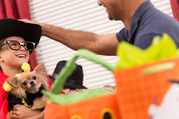 Cute little Latin descent boy dresses up for Halloween party or to go trick or treat with the help of his dad.  The boy holds his pet dog, who is dressed in a bee costume.  The pair have lots of fun choosing costumes at home family room setting.