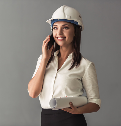 Beautiful young business woman in suit and protective helmet is talking on the mobile phone, holding rolled paper and smiling, on gray background