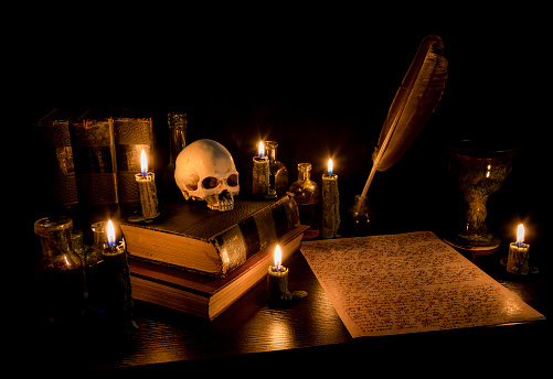 Wizard's Desk. A desk lit by candle light. A human skull, old books, a goblet, and potion bottles are present. Desk has parchment paper with arcane writing. Feather pen in inkwell. Focus on skull.