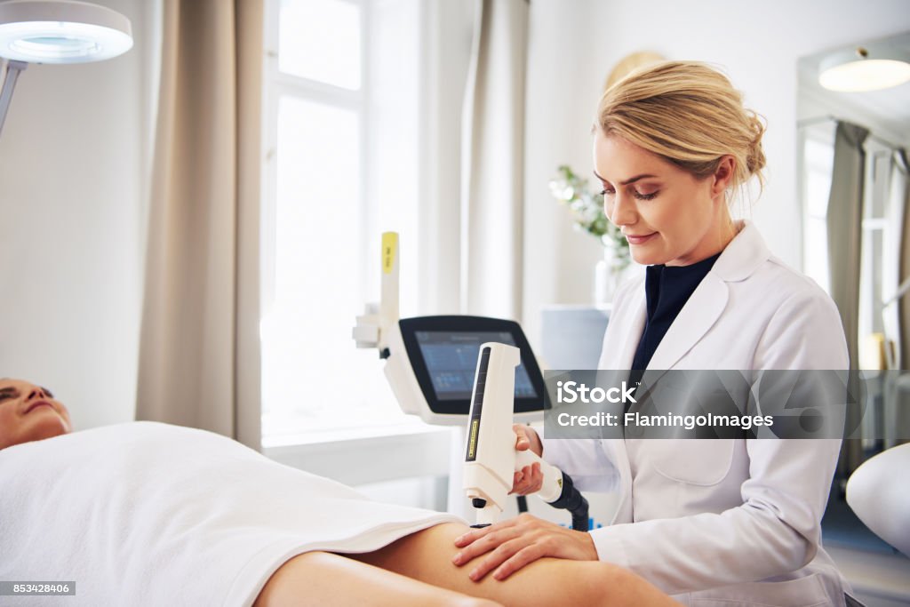 Clinic technician performing laser hair removal on a client's leg Female technician performing a laser hair removal procedure on the legs of a young woman lying on a table in a beauty clinic Dermatology Stock Photo