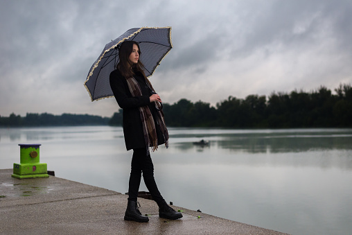 Beautiful young woman in black standing on river dock beside a river on the rainy autumn day.