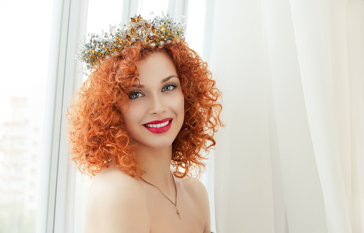 Happy bride. Beautiful red head curly woman with crown fashion model person girl looking at you camera smiling at boyfriend groom isolated  near window indoor white curtains background. Miss sunshine
