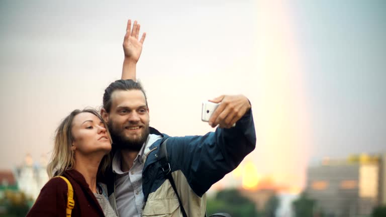 Happy couple standing on the city centre on sunset, taking selfie photos with rainbow on the background on smartphone