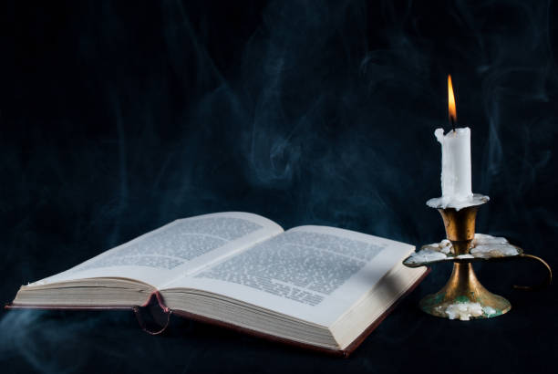 istock Old open book and candle in candlestick and smoke on dark black background 853547612