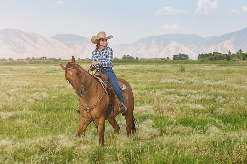 Confident cowgirl riding with her horse through the ranch grassland, looking over. Real People Farmer Portrait. Utah, USA.