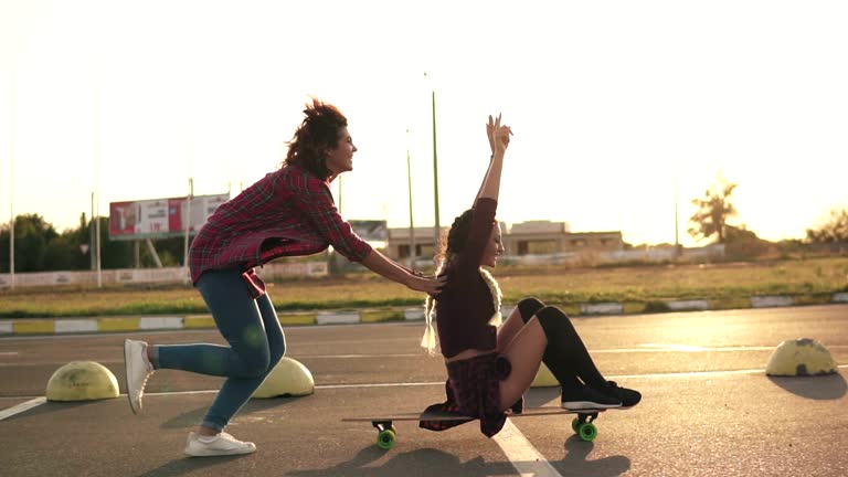 Side view of a woman with her hands raised up sitting on a longboard while her friend is pushing her behind and running during sunset. Enjoying life. Lens flare. Slowmotion shot