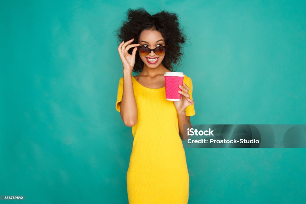 Happy girl drinking coffe at studio background Curly smiling girl in bright yellow dress and sunglasses with take away coffee cup. Young woman at azur studio background looking cunningly from under her dark glasses, copy space Coffee - Drink Stock Photo