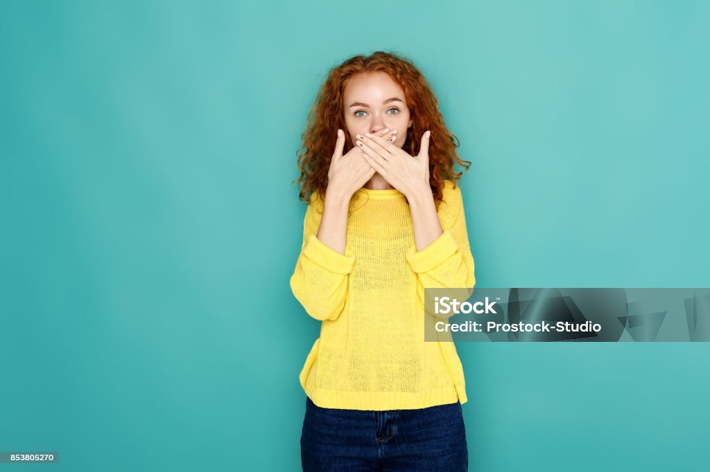 Scared woman covering mouth with hands Keep silence. Redhead woman covering mouth with hands while posing to camera on blue studio background. Girl close lips with palms, speak no evil concept 20-29 Years Stock Photo